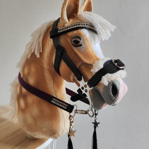 May include: A tan plush hobby horse head with a white mane and forelock. It has a black bridle with silver details and a purple strap. The horse features a decorative headstall with rhinestones and black tassels with star charms.