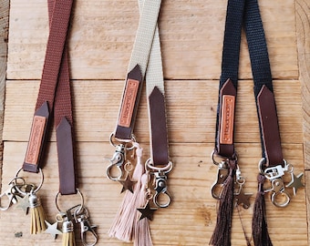 May include: Three lanyards in rust, beige, and black fabric with brown leather accents. Each features a tag embossed with the word handmade, a silver-toned metal lobster clasp, a hanging star charm, and a matching thread tassel at the base.