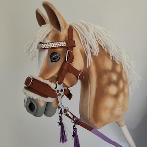 Op de afbeelding: Een pluche stokpaardenkop in tinten bruin en beige, met een witte manen en een bruine teugel versierd met strass. Het paard heeft een grijze snuit en een paarse teugel met kwastjes.