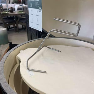 May include: A stainless steel grill tool rests on a white ceramic heat deflector inside a charcoal grill. The grill has a dark exterior and a light interior. A price tag is visible in the background.