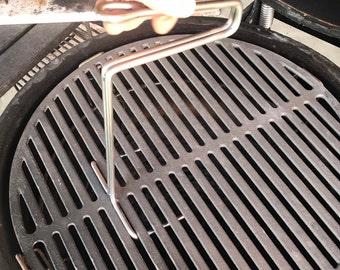 May include: A close-up of a silver grill grate lifter being used to lift a cast iron grill grate. The grate is in a large, round, black grill.