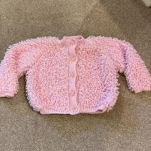 May include: A light pink long-sleeve cardigan featuring a textured loop stitch pattern throughout the body and sleeves. The garment has a ribbed crew neckline, a vertical ribbed front placket with four matching pink buttons, and ribbed cuffs.
