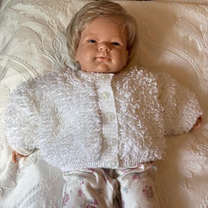 May include: A doll with blonde hair wearing a white long-sleeved cardigan with a textured loop stitch pattern and four clear buttons down the front. The doll is also wearing white trousers featuring a small pink and green floral pattern throughout.