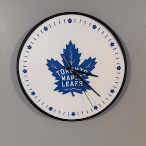 Peut inclure: Une horloge Toronto Maple Leafs blanche et bleue avec un cadre noir. L'horloge présente une feuille d'érable bleue avec le nom de l'équipe imprimé en dessous.
