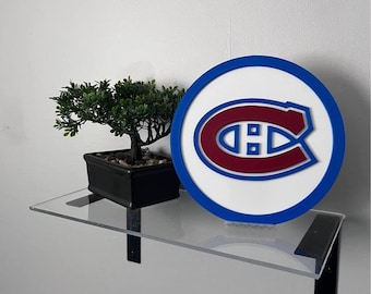 Könnte beinhalten: Ein rundes Schild mit dem Logo der Montreal Canadiens in Rot, Weiß und Blau. Das Schild steht auf einem transparenten Regal zusammen mit einem kleinen Bonsai-Baum in einem schwarzen Topf. Das Regal wird von schwarzen Metallhalterungen getragen.
