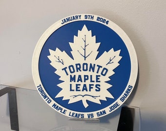 Könnte beinhalten: Ein blaues und weißes rundes Schild mit einem weißen Ahornblatt und dem Text "Toronto Maple Leafs" und "January 9th 2024". Das Schild hat auch den Text "Toronto Maple Leafs vs San Jose Sharks" am Rand.