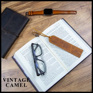 May include: A brown leather bookmark with a frayed edge, a book with reading glasses on top, and a brown leather wristwatch with a black face on a wooden surface.