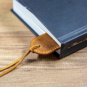 May include: A brown leather bookmark with a stitched edge is tucked into the corner of a dark blue hardcover book. The book has gold lettering on the spine that reads "Macabre"