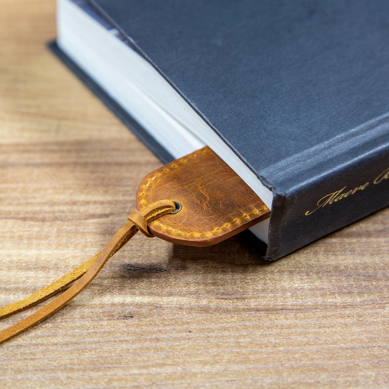 May include: A brown leather bookmark with a stitched edge is tucked into the corner of a dark blue hardcover book. The book has gold lettering on the spine that reads "Macabre"