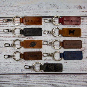 May include: A set of ten leather keychains with different designs and engravings. The keychains are brown, black, red, blue, and gray. The engravings include "Dad's Keychain", "Mom's Keyring", "I Love You", "Happy Birthday", "Brittany & Andrew", "23.01.1987", a football, a dog, and a yin yang symbol.