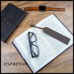 May include: A brown leather bookmark with a frayed edge, laying on an open book with a pair of black reading glasses. The book is open to a page with text. The bookmark is labeled "ESPRESSO".