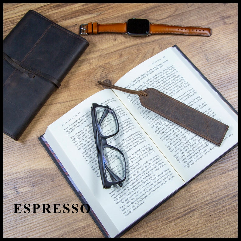 May include: A brown leather bookmark with a frayed edge, laying on an open book with a pair of black reading glasses. The book is open to a page with text. The bookmark is labeled "ESPRESSO".