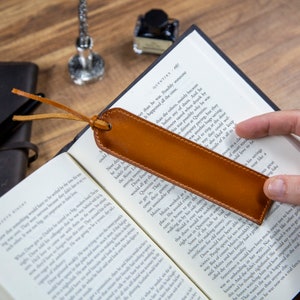 May include: A brown leather bookmark with a tan leather tassel. The bookmark is lying on an open book with text. The bookmark is a useful tool for keeping your place in a book.