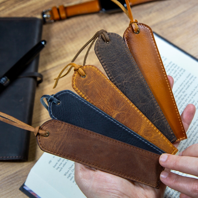 May include: Four leather bookmarks in different colors, including brown, dark brown, blue, and a lighter brown. The bookmarks are arranged in a fan shape and are held in a hand.