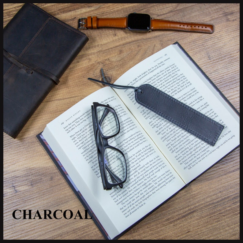 May include: A charcoal leather bookmark with a stitched edge, laying on an open book with a pair of reading glasses and a brown leather wristwatch.