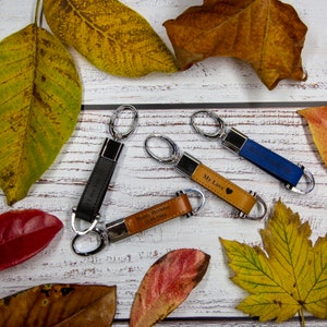 May include: Four leather keychains with different engravings. The keychains are brown, black, and blue. The keychains are on a white wooden surface with fall leaves scattered around them. The engravings on the keychains read "I Love You", "My Love", "Happy Birthday 12.06.1984", and "Best Dad Ever".