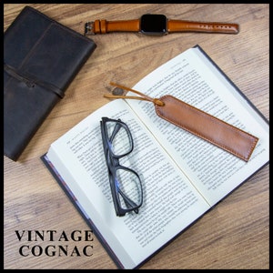 May include: A brown leather bookmark with a tassel, a pair of black reading glasses, a brown leather journal, and a brown leather watch band with a silver watch face on a wooden surface. The text "VINTAGE COGNAC" is visible in the lower left corner of the image.