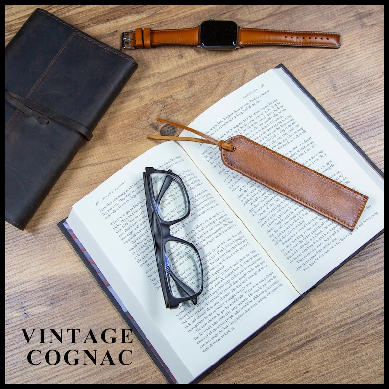 May include: A brown leather bookmark with a tassel, a pair of black reading glasses, a brown leather journal, and a brown leather watch band with a silver watch face on a wooden surface. The text "VINTAGE COGNAC" is visible in the lower left corner of the image.