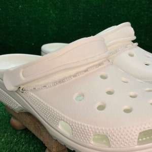 May include: White clogs with a beaded strap. The clogs have a textured surface and multiple ventilation holes. The strap is adorned with small, silver-colored beads. The clogs are placed on a wooden surface against a green background.
