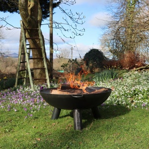 May include: A black fire pit with burning logs, set on a lawn with a wooden ladder and spring flowers. The flames are orange and yellow. The fire pit has three legs and a round bowl shape, perfect for outdoor gatherings.
