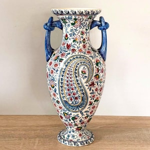 Op de afbeelding: Een keramische vaas in amfoorstijl met een witte basis, voorzien van complexe bloemmotieven in blauw, rood en groen en een centraal paisley-patroon. De vaas heeft twee effen blauwe handvatten met een knoopontwerp en een uitlopende rand.