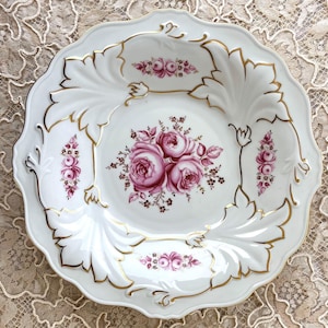 May include: A white porcelain serving bowl with a gold rim and intricate floral designs in shades of pink and purple. The bowl has a scalloped edge and a raised center.