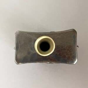 Op de afbeelding: Bovenaanzicht van een zilveren metalen vaas met een ronde opening. De vaas heeft een licht gebogen rechthoekige vorm en een getextureerd oppervlak.