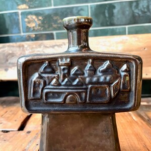 Op de afbeelding: Een donkerbruine keramische fles met een reli&euml;fdesign van een middeleeuwse stad. De fles heeft een vierkante basis en een smalle hals.