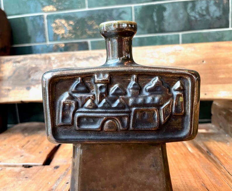 Op de afbeelding: Een donkerbruine keramische fles met een reli&euml;fdesign van een middeleeuwse stad. De fles heeft een vierkante basis en een smalle hals.