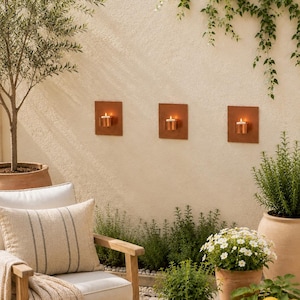May include: Three square copper wall sconces mounted on a beige wall, each holding a lit tea light candle. A matching copper candle holder sits on a wooden table alongside a wooden bowl filled with yellow lemons and potted green plants.