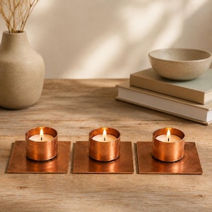 May include: Three cylindrical copper tea light candle holders, each mounted on a square copper base. Each holder contains a small white wax candle with a lit flame. The metallic surfaces have a warm, reflective finish and a smooth texture.