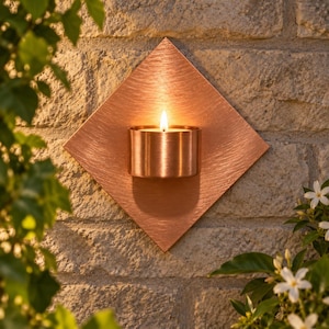 May include: A diamond-shaped copper wall sconce featuring a brushed metal finish and a central cylindrical holder containing a lit tea light candle. The fixture is mounted against a textured stone wall, casting a warm glow from the flame.