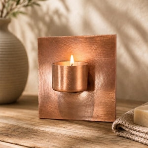 May include: A square copper candle holder with a brushed metallic finish and a central cylindrical cup. A small tea light candle is lit inside the cup, emitting a warm glow against the flat, reflective copper surface of the square backing.