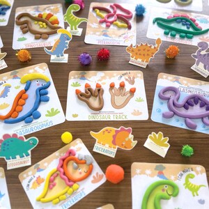 K&ouml;nnte beinhalten: Eine Sammlung von Dinosaurier-Knetkarten mit bunten Tonfiguren verschiedener pr&auml;historischer Kreaturen wie Parasaurolophus, Elasmosaurus und Triceratops sowie Abdr&uuml;cken von Dinosaurierspuren auf wei&szlig;en Karten.