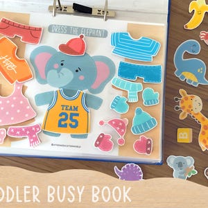 Op de afbeelding: Een activiteitenboek voor peuters met een centrale olifant en de tekst DRESS THE ELEPHANT. De set bevat diverse afneembare kledingstukken zoals shirts, mutsen en shorts, naast extra stickers van dieren en voedsel.