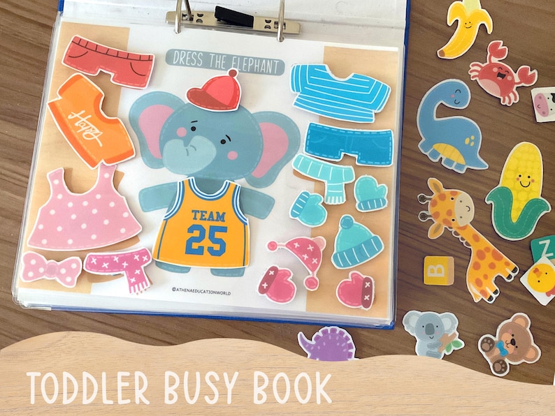 Op de afbeelding: Een activiteitenboek voor peuters met een centrale olifant en de tekst DRESS THE ELEPHANT. De set bevat diverse afneembare kledingstukken zoals shirts, mutsen en shorts, naast extra stickers van dieren en voedsel.