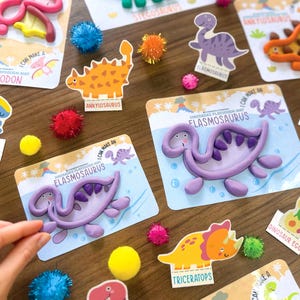 K&ouml;nnte beinhalten: Eine Sammlung von Knetmatten mit Dinosaurier-Motiven, die Illustrationen von Elasmosaurus, Pteranodon, Ankylosaurus, Parasaurolophus und Triceratops zeigen, erg&auml;nzt durch bunte Pompons und lila Knete in Form von Dinosauriern.