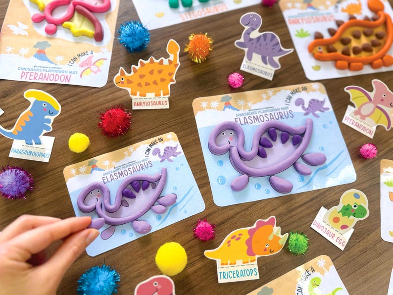 K&ouml;nnte beinhalten: Eine Sammlung von Knetmatten mit Dinosaurier-Motiven, die Illustrationen von Elasmosaurus, Pteranodon, Ankylosaurus, Parasaurolophus und Triceratops zeigen, erg&auml;nzt durch bunte Pompons und lila Knete in Form von Dinosauriern.