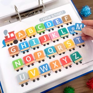 Op de afbeelding: Een educatieve map-pagina getiteld I KNOW ALPHABET met een kleurrijke treinafbeelding met rails. Vierkante tegels met witte letters van A tot Z zijn in rijen op de treinwagons geplaatst. De letter M wordt op de pagina geplaatst.
