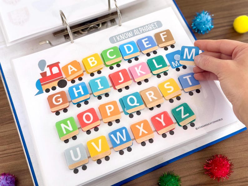 Op de afbeelding: Een educatieve map-pagina getiteld I KNOW ALPHABET met een kleurrijke treinafbeelding met rails. Vierkante tegels met witte letters van A tot Z zijn in rijen op de treinwagons geplaatst. De letter M wordt op de pagina geplaatst.