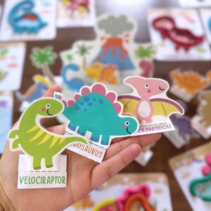 K&ouml;nnte beinhalten: Drei ausgestanzte Dinosaurierfiguren aus Papier, bestehend aus einem gr&uuml;nen Velociraptor, einem blaugr&uuml;nen Stegosaurus und einem rosa Pteranodon. Jede Figur hat eine wei&szlig;e Bodenlasche mit dem Namen in schwarzer Schrift zum Aufstellen.