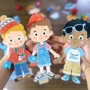 Pode incluir: Tr&ecirc;s figuras de papel com roupas diversas. A figura da esquerda usa um gorro de inverno vermelho, um su&eacute;ter com um boneco de neve e luvas azuis. A figura central usa um bon&eacute; vermelho e jardineiras azuis. A figura da direita usa &oacute;culos de sol e uma camiseta com o texto Summer Vibes.
