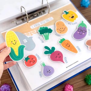 Op de afbeelding: Een educatieve map-pagina getiteld I KNOW VEGETABLES met kleurrijke, cartoonachtige groentestickers met lachende gezichten, waaronder ma&iuml;s, broccoli, aardappel, paprika, aubergine, tomaat, radijs, wortel, champignon en ui met bijbehorende labels.