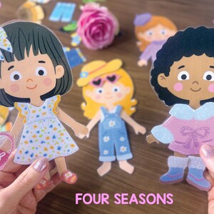 Pode incluir: Tr&ecirc;s bonecas de papel com penteados e roupas distintas, incluindo uma menina com vestido floral e la&ccedil;o, uma menina com chap&eacute;u amarelo e jardineiras jeans, e uma menina de cabelo encaracolado com casaco rosa, com o texto FOUR SEASONS.
