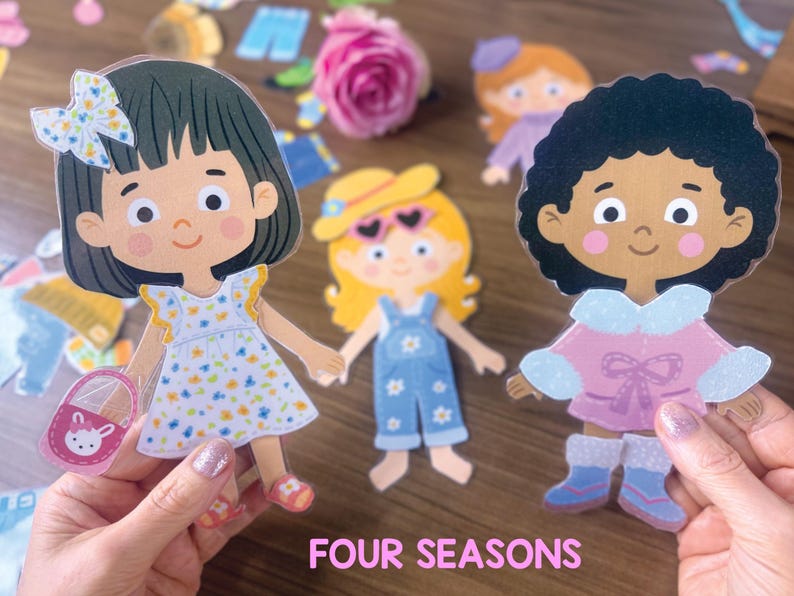 Pode incluir: Tr&ecirc;s bonecas de papel com penteados e roupas distintas, incluindo uma menina com vestido floral e la&ccedil;o, uma menina com chap&eacute;u amarelo e jardineiras jeans, e uma menina de cabelo encaracolado com casaco rosa, com o texto FOUR SEASONS.
