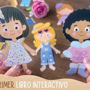 May include: Interactive paper dolls with outfits. The dolls have dark hair, blonde hair, and curly black hair. The dolls are wearing a dress, overalls, and a winter coat. The text "MI PRIMER LIBRO INTERACTIVO" is at the bottom.