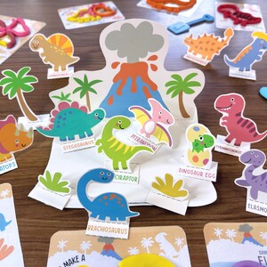 K&ouml;nnte beinhalten: Ein Papierbastelset mit einem zentralen Vulkan-Aufsteller und verschiedenen Dinosaurier-Ausschnitten, darunter Triceratops, Stegosaurus, Pteranodon, Tyrannosaurus, Brachiosaurus und Elasmosaurus, die jeweils mit ihrem Namen auf einem Sockel stehen.