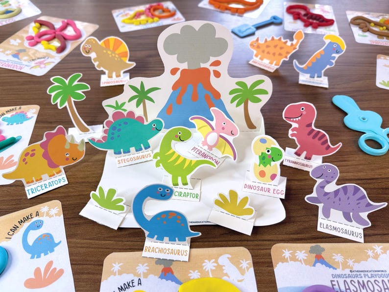K&ouml;nnte beinhalten: Ein Papierbastelset mit einem zentralen Vulkan-Aufsteller und verschiedenen Dinosaurier-Ausschnitten, darunter Triceratops, Stegosaurus, Pteranodon, Tyrannosaurus, Brachiosaurus und Elasmosaurus, die jeweils mit ihrem Namen auf einem Sockel stehen.