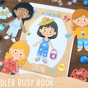 Op de afbeelding: Een papieren poppenset getiteld Dress The Girl Spring met een centraal figuur in een blauwe tuinbroek met madeliefjesprint, een strooien hoed en een roze tas, omringd door extra papieren poppen en diverse kledingstukken zoals truien, broeken en sokken.