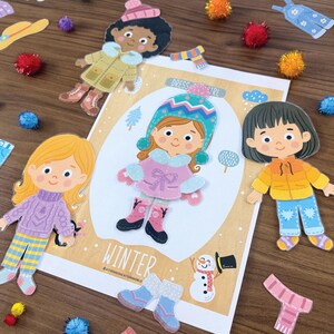 Pode incluir: Um conjunto de bonecas de papel com um cart&atilde;o central escrito WINTER, mostrando uma personagem com casaco rosa e gorro azul. Em torno do cart&atilde;o, h&aacute; duas bonecas adicionais, v&aacute;rias pe&ccedil;as de roupa como camisolas e cal&ccedil;as, e pompons coloridos.